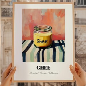 Op de afbeelding: Een ingelijste print met een pot ghee, met het woord "Ghee" in geel op de pot. De print is geplaatst tegen een kleurrijke abstracte achtergrond en een gestreepte tafel. De tekst "Essential Flavors Collection" staat onder het woord "Ghee".