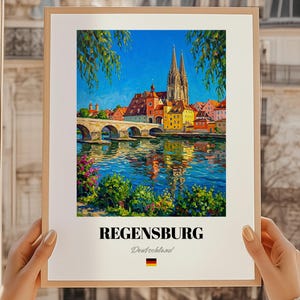 Könnte beinhalten: Ein gerahmter Druck von Regensburg, Deutschland, mit einer lebendigen Stadtlandschaft und einer Brücke über einem Fluss. Das Kunstwerk zeigt farbenfrohe Gebäude, eine Kathedrale mit Doppeltürmen und üppiges Grün unter einem strahlend blauen Himmel. Der Druck enthält den Namen der Stadt und die deutsche Flagge.