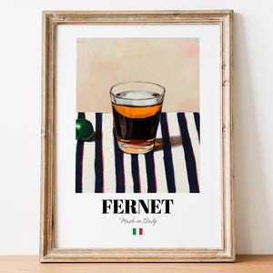 May include: A glass of amber-colored liquid sits on a white and blue striped surface. The glass is clear and has a rounded bottom. The liquid is a dark amber color and has a small amount of foam on top. The surface is a white and blue striped fabric. The text "FERNET Made in Italy" is printed below the glass. A small green lime is visible in the background.