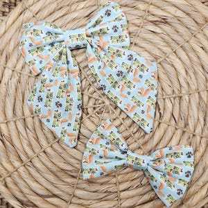 May include: Three light blue fabric bows with a repeating pattern of orange squirrels, paw prints, and the text "SQUIRREL PATROL". The bows are arranged on a woven, light brown surface.