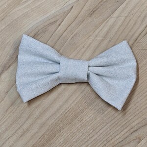 May include: A silver glitter bow tie, a formal accessory, lies on a wooden surface. The bow tie is made of a shimmering fabric, with a classic bow shape. The bow tie is a light silver color.