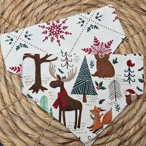 May include: Two cream-colored dog bandanas with a winter forest pattern. One features a snowflake design, while the other showcases a moose with a red scarf, a fox on a sled, and a bear. The bandanas are made of soft fabric.