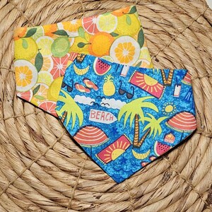 May include: Two fabric bandanas with a summer theme. One bandana is yellow with a citrus fruit pattern. The other bandana is blue with a palm tree, watermelon, and sun pattern. The text "Beach" is printed on the blue bandana.