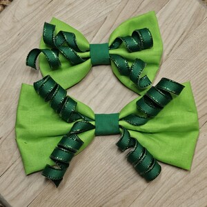 May include: Two bright green fabric hair bows with dark green ribbon accents. The bows have a classic bow shape with a center band and decorative, spiraling ribbon curls. The bows are set against a light wood grain background.