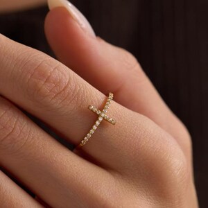 May include: A gold ring with a cross design, set with small, sparkling stones. The ring is worn on a finger, highlighting its delicate and elegant appearance. The ring is a piece of jewellery. The ring is a piece of jewellery.