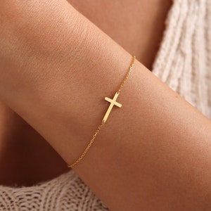 May include: A delicate gold-colored bracelet featuring a sideways cross pendant. The cross is attached to a fine chain, creating a minimalist and elegant design. The bracelet is worn on a wrist, showcasing its simple yet meaningful aesthetic.