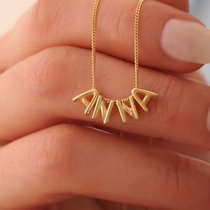 Könnte beinhalten: Eine goldfarbene Halskette mit einer feinen Kette und einzelnen goldfarbenen Buchstabenanhängern, die den Namen ANNA bilden. Die Buchstaben hängen an der Kette und erzeugen ein horizontales Namensschild-Design mit einer polierten Oberfläche.