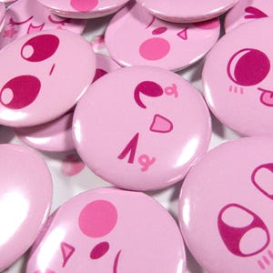 May include: A collection of pink, round buttons with various cartoon faces in shades of pink. The buttons feature different expressions, including winking, smiling, and sticking out the tongue. The buttons are scattered across the surface.