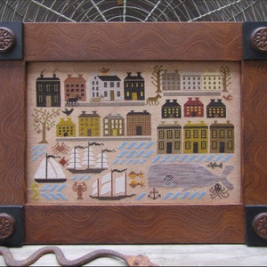 Op de afbeelding: Een ingelijst borduurwerk met kruissteek, dat een kustplaats voorstelt met huizen, schepen, een walvis en andere nautische details. De lijst is van bruin hout met decoratieve hoekaccenten.