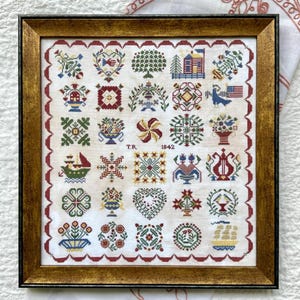 May include: Framed cross-stitch sampler with a gold-colored frame. The sampler features a grid of colorful embroidered motifs, including hearts, flowers, trees, and geometric patterns. The year 1842 is stitched into the design.