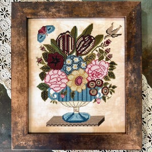 May include: A framed cross-stitch embroidery depicting a blue and white striped vase filled with a colorful bouquet of flowers, including pink roses, yellow daisies, and red poppies. The vase is on a small pedestal and there are butterflies and birds in the background.