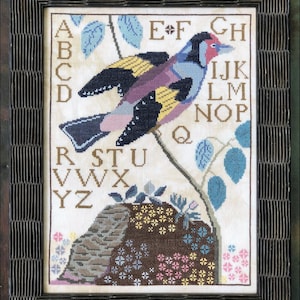 Op de afbeelding: Borduurwerk met kruissteek van een kleurrijke vogel die op een tak zit met groene bladeren. Het alfabet is rond de vogel geborduurd in een vintage lettertype. De achtergrond is een crèmekleurige stof.