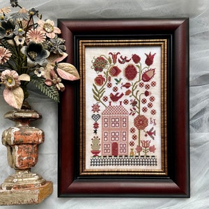 May include: A framed cross-stitch embroidery depicting a pink house with a red roof, surrounded by a garden with red flowers, green leaves, and small birds. The house has a red door and six windows. The embroidery is in a dark brown frame with a gold inner border.