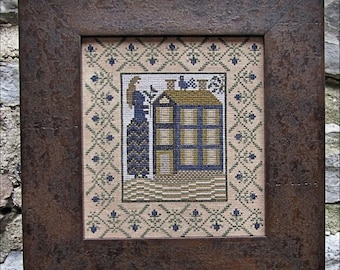 May include: A framed cross-stitch embroidery featuring a woman in a blue dress standing in front of a small house with a brown roof and blue windows. The background is a white fabric with a repeating pattern of green leaves and blue flowers.