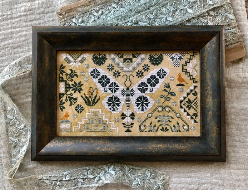 Pu&ograve; includere: Un ricamo a punto croce incorniciato che presenta una farfalla in bianco e nero con motivi geometrici. Lo sfondo &egrave; un tessuto beige con vari disegni floreali e geometrici in tonalit&agrave; di verde, marrone e bianco.