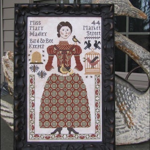 May include: A framed cross-stitch embroidery featuring a woman in a red dress and a birdcage with birds. The text reads "Miss Mary Madley Bird & Bee Keeper 44 Market Street".