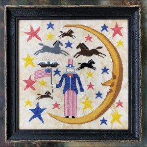 May include: A cross-stitch embroidery design featuring Uncle Sam standing on a crescent moon with stars and horses in the background. The design is framed in a black wood frame.