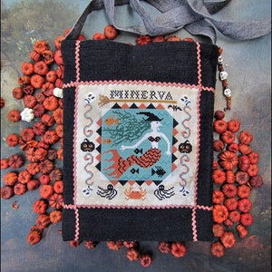 May include: Black crossbody bag with a stitched design featuring a mermaid, fish, and the name "MINERVA". The bag has a gray strap and is surrounded by small orange pumpkins. The design includes a witch hat, pumpkins, and a crab.