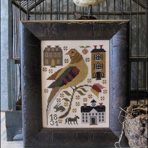 Op de afbeelding: Een ingelijst borduurwerk met kruissteek, met een vogel die op een tak zit met huizen en bloemen op de achtergrond. Het borduurwerk is in een donkerbruin frame met een verweerde afwerking. De nummers "18 31" zijn in de linkerbenedenhoek geborduurd.