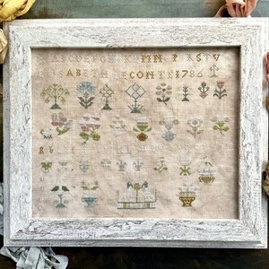 May include: A framed cross-stitch sampler with a white frame and a beige linen background. The sampler features a variety of floral motifs, including flowers, leaves, and stems. The sampler also includes the alphabet, numbers, and the year 1786.