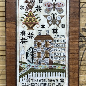 May include: A framed cross-stitch sampler featuring a white house with a water wheel, a bird, butterflies, and a fence. The sampler is signed "The Mill House Catherine Miller in 1887".