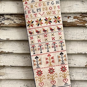 May include: A beige linen Christmas stocking featuring intricate cross-stitch embroidery in red, black, and gold. The design includes multiple alphabets, rows of stylised floral motifs, birds, hearts, and a peacock figure, with the year 1821 stitched at the bottom.