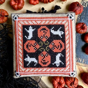 Op de afbeelding: Een vierkante, kruissteek Halloween-decoratie met oranje pompoenen en witte geesten op een zwarte achtergrond. Het ontwerp is omlijst met een cr&egrave;me- en oranje rand. Kleine, decoratieve pompoenen omringen het gestikte stuk.