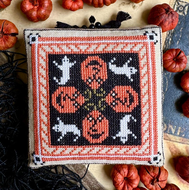 Op de afbeelding: Een vierkante, kruissteek Halloween-decoratie met oranje pompoenen en witte geesten op een zwarte achtergrond. Het ontwerp is omlijst met een cr&egrave;me- en oranje rand. Kleine, decoratieve pompoenen omringen het gestikte stuk.