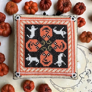 Peut inclure: Une décoration carrée de Halloween brodée au point de croix, avec des citrouilles orange et des fantômes blancs sur fond noir. Le motif est encadré de bordures orange, crème et noires. De petites citrouilles oranges entourent la décoration.