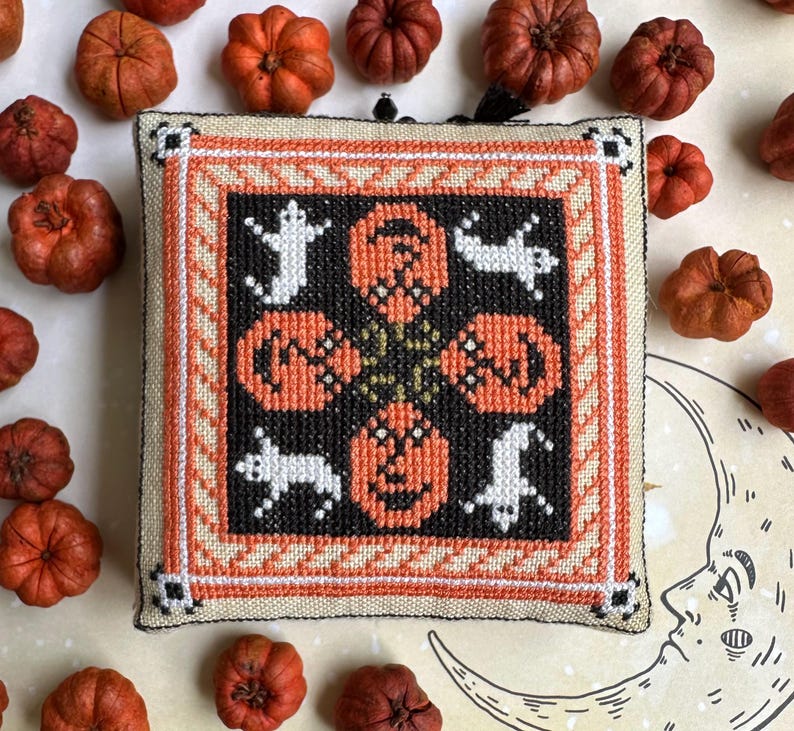 Op de afbeelding: Een vierkante, kruissteek Halloween-decoratie met oranje pompoenen en witte spookfiguren op een zwarte achtergrond. Het ontwerp is omlijst met oranje, cr&egrave;me en zwarte randen. Kleine, oranje pompoenen omringen de decoratie.