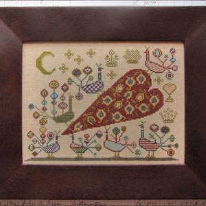 Op de afbeelding: Een ingelijste kruissteek sampler met een groot hartvormig ontwerp in rood en goud. Het ontwerp bevat gestileerde vogels, bloemen en een maan op een beige achtergrond. De lijst is gemaakt van donkerbruin hout.