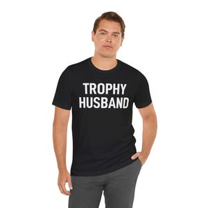 May include: Black crew neck t-shirt with the words "TROPHY HUSBAND" printed in white block letters. The shirt is worn by a person in gray pants. The background is white.