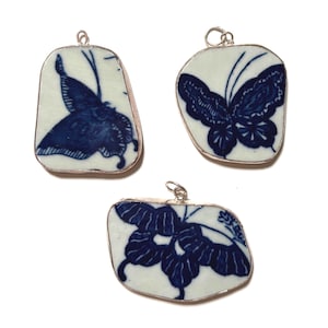 May include: Three irregular, flat ceramic pendants featuring blue and white butterfly patterns. Each piece is encased in a thin silver-toned metal border and includes a small circular metal loop at the top for attachment to a necklace or chain.