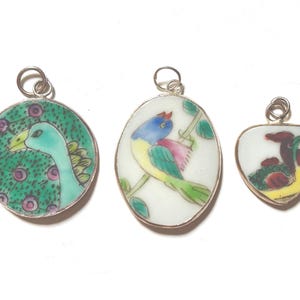 May include: Three pendants with silver-toned metal rims and jump rings. The circular pendant features a green bird illustration. The oval pendant shows a multi-colored bird on a branch. The heart-shaped pendant depicts a small bird with red accents.