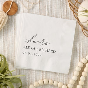 May include: White paper cocktail napkin with black calligraphy that reads "cheers" followed by the names "Alexa + Richard" and the date "06.01.2024".