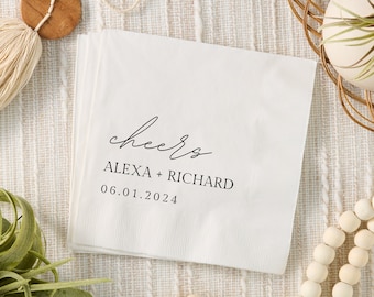 May include: White paper cocktail napkin with black calligraphy that reads "cheers" followed by the names "Alexa + Richard" and the date "06.01.2024".