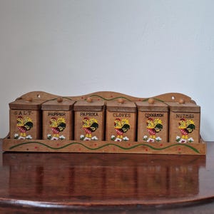 Pode incluir: Um porta-temperos de madeira com seis recipientes retangulares. Cada recipiente é rotulado com o nome de uma especiaria e apresenta uma ilustração colorida de um galo. O suporte tem uma borda decorativa ondulada e um design pintado em verde e vermelho.