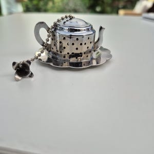 May include: A silver-coloured metal tea infuser shaped like a miniature teapot with a perforated body, a handle, and a spout. It rests on a matching scalloped saucer and includes a long metal ball chain with a small decorative charm at the end.