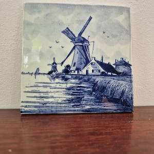 Può includere: Piastrella quadrata in ceramica con un'illustrazione blu e bianca di una scena di mulino a vento olandese. L'opera raffigura mulini a vento, case, acqua e uccelli contro un cielo nuvoloso. La piastrella è esposta su una superficie di legno.