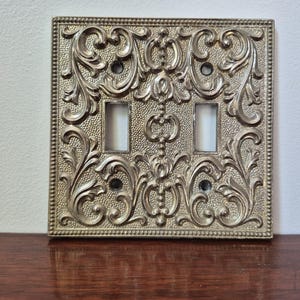 May include: An antique-style light switch cover with an ornate, square design. The metallic cover features a detailed, raised floral and scrollwork pattern. It has two rectangular switch openings and four screw holes, showcasing a vintage aesthetic.