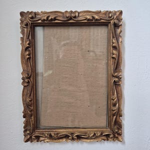 May include: An antique-style rectangular picture frame with a gold finish. The frame has elaborate carved floral and scrollwork patterns. The glass-covered space within the frame is empty, ready for a picture. The frame is mounted on a white wall.