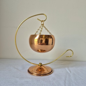 May include: A polished copper-coloured bowl suspended by a gold-toned metal chain from a curved gold-toned metal stand. The stand features a circular base that matches the copper finish of the bowl, creating a reflective and metallic display piece.