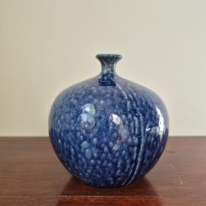 Peut inclure: Un vase rond en céramique bleue avec une surface texturée et mouchetée. Le vase a un col étroit et un corps bulbeux. L'émail bleu foncé a des accents bleus clairs et blancs. Le vase est posé sur une surface en bois foncé.