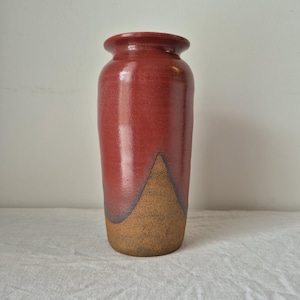 May include: A cylindrical ceramic vase featuring a glossy, deep red glaze on the upper portion and a textured, matte brown finish on the lower section. The vase has a distinct flared rim and a smooth transition between the two contrasting colours.