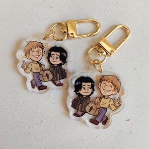 May include: Two clear acrylic keychains featuring a cartoon illustration of two characters walking together. One character has light brown hair and a yellow sweater, while the other has black hair and a dark jacket. Both carry bags and gold clasps.
