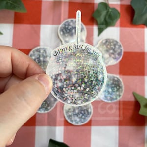 Könnte beinhalten: Ein runder Disco-Kugel-Aufkleber mit holografischer Oberfläche, die Licht in verschiedenen Farben reflektiert. Der Aufkleber trägt den Schriftzug "shining just for you" in dunkler Schrift in der Mitte und hat eine kleine vertikale Lasche oben.