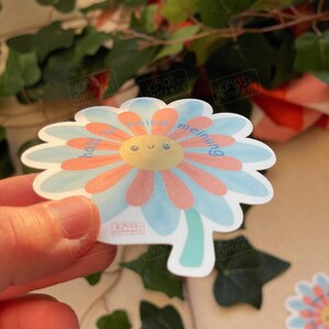 Puede incluir: Una pegatina con forma de margarita con una cara sonriente en el centro, rodeada de p&eacute;talos naranjas y celeste. La pegatina tiene las palabras "keine meinung" en azul. La pegatina est&aacute; sujeta por una mano.