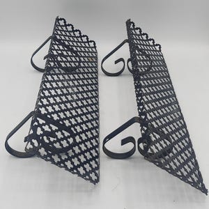 May include: Two black metal wall shelves featuring a perforated lattice pattern with a repeating cross design. Each shelf is supported by two curved, scroll-shaped brackets that extend from the back, providing a stable mounting structure.