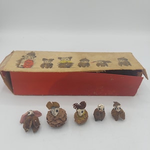 May include: Five small figurines crafted from natural materials like pinecones and seed pods, each featuring a painted face. The figurines are displayed in front of a rectangular cardboard box with a red base and a tan lid featuring illustrations of the figures.