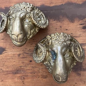 Puede incluir: Dos esculturas de cabezas de carnero en metal de color dorado con cuernos curvados detallados y patrones de lana texturizados. Cada cabeza de carnero está moldeada con una expresión facial realista, incluyendo ojos, hocico y orejas bien definidos.